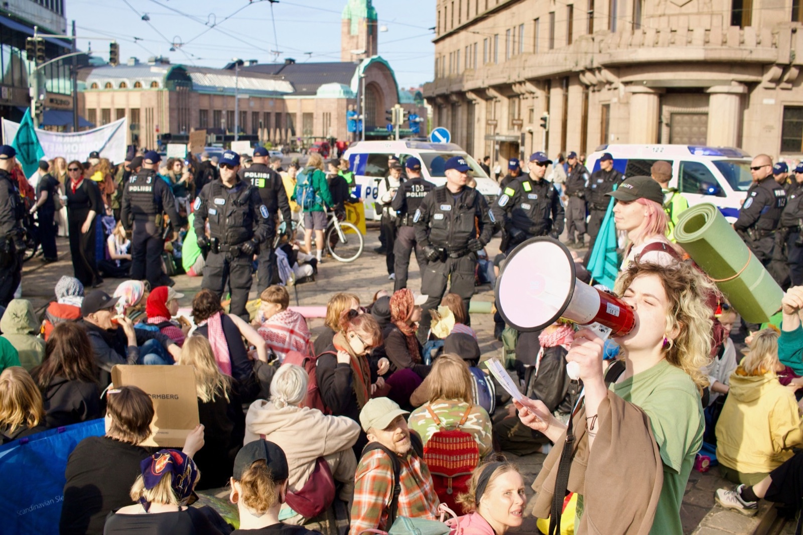 Mielenosoittajat istuvat Mannerheimintiellä Helsingin rautatieaseman edessä, poliiseja heidän takanaan. Etualalla nuori nainen puhuu megafoniin.