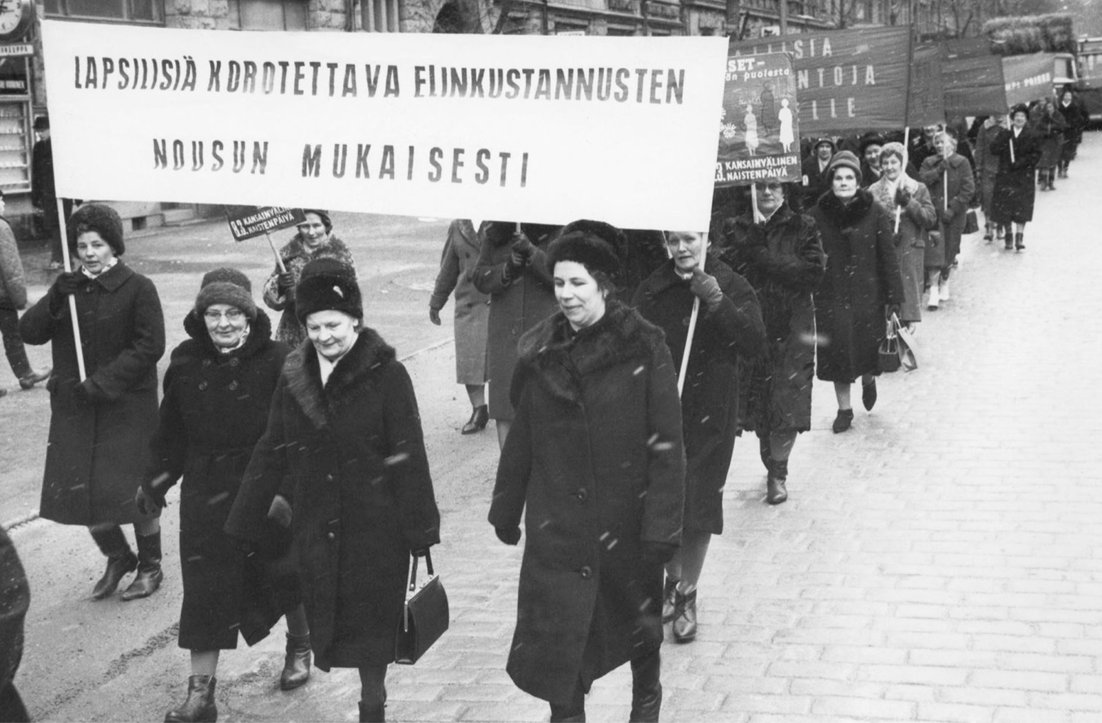 Mustavalkoinen kuva: talvitakkeihin pukeutuneet naiset marssivat kadulla banderollin takana. Banderollissa lukee: Lapsilisä korotettava elinkustannusten nousun mukaisesti.